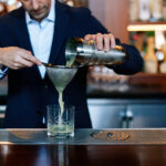 bartender pouring a light green snap pea cocktail through a sieve and into a rocks glass